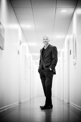 Black and white image of bald man in a black jacket and pants standing in a bright white corridor