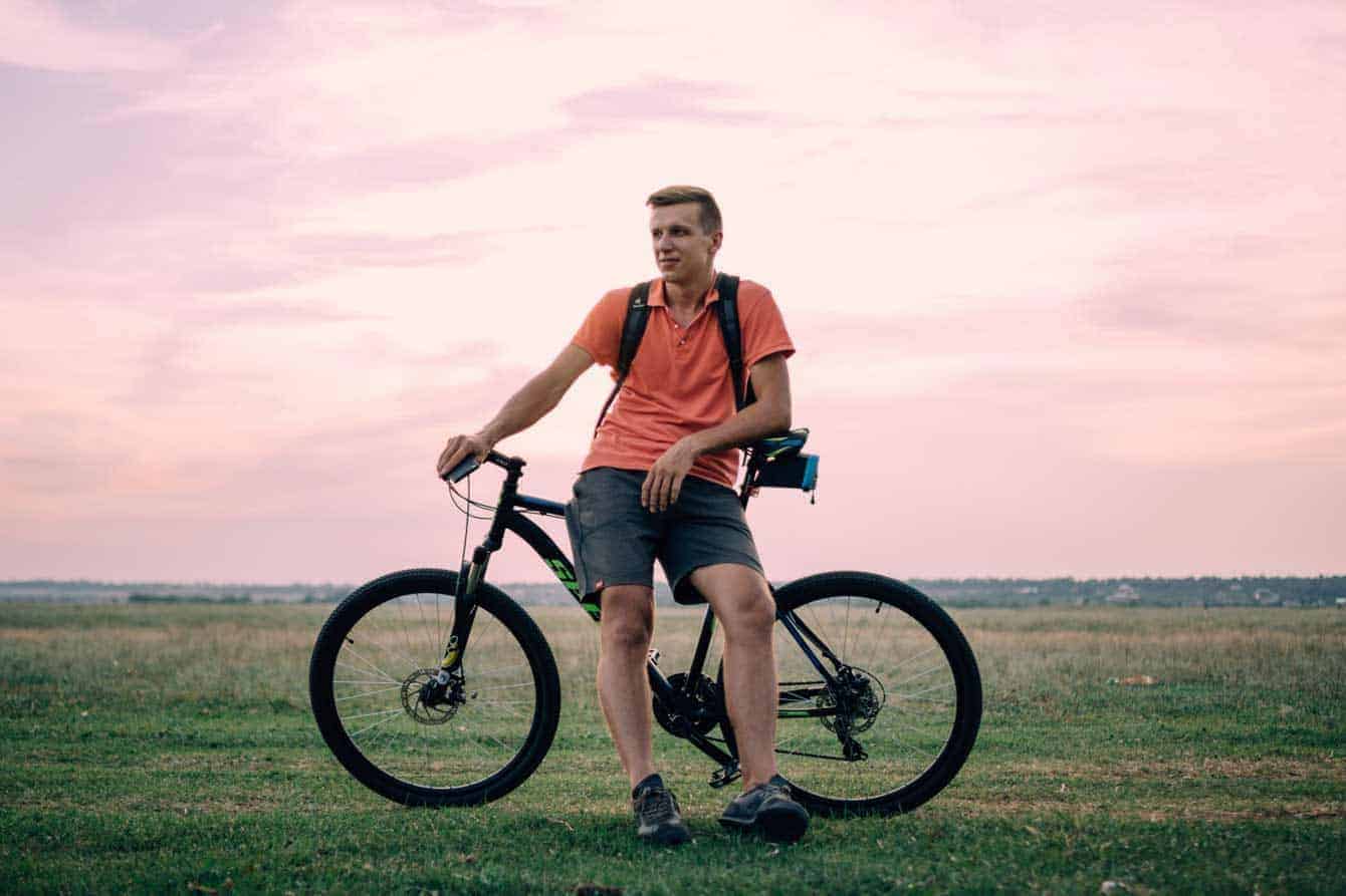 Man in an orange shirt and blue shorts leaning against a bike in a field at sunset