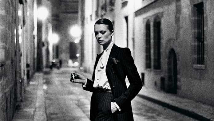 A striking black-and-white photo capturing a poised model in a tailored suit, standing alone in a dimly lit urban alley. Strong contrasts and vintage ambiance evoke timeless elegance and.