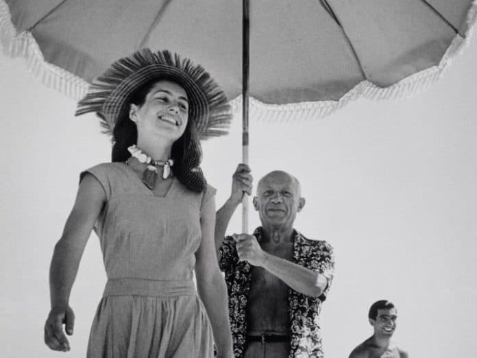 Black and white photo of a smiling woman in a sun hat and dress, walking under a large umbrella held by a man, with two other figures in the background, capturing a relaxed beach.