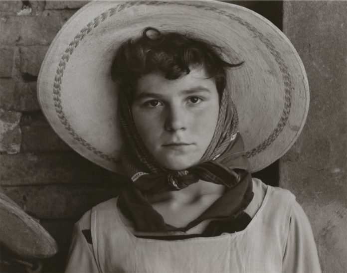 A striking portrait of a young person wearing a large straw hat and scarf, set against a textured background, capturing emotion and authenticity in black and white.