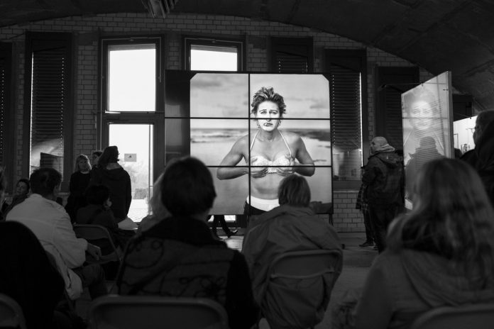 A dynamic black and white scene featuring an audience captivated by a large screen displaying a striking portrait. The setting is filled with natural light and artistic tension, highlighting powerful.