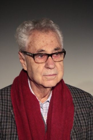 A distinguished elderly man with gray hair and glasses, wearing a plaid coat and a vibrant red scarf, is captured against a soft, neutral background. This image represents wisdom in.