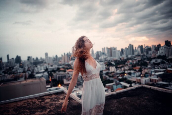 Woman in white dress standing with her arms back and face to a dramatic, cloudy sky while standing on an apartment building rooftop.