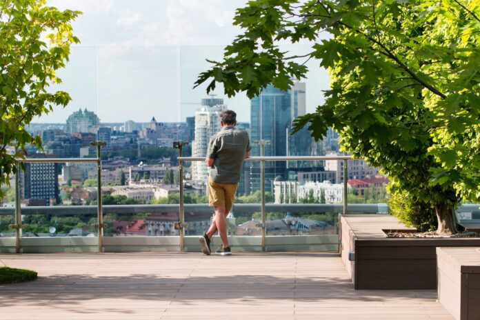 Man standing at the railing of a rooftop garden looking out over city, taken from behind him.