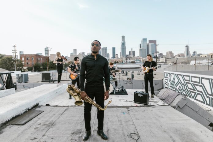 Band photoshoot with saxophonist standing far in front of his band on warehouse roof with cityscape in background.