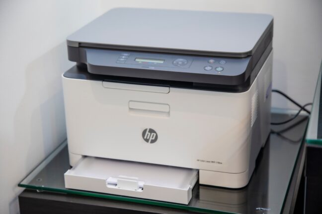 Blue and white HP color laser printer in corner of a room on a glass table with printer tray open.