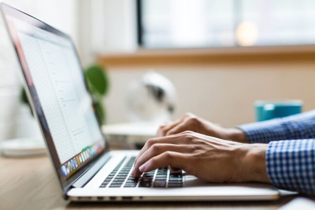 Hands typing on an Apple MacBook laptop with a coffee cup and a desk plant in the background