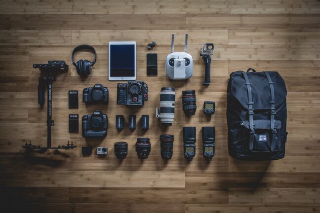 Cameras, lenses, a backpack, headphones, a tablet, and other photo and video electronics on a wooden floor