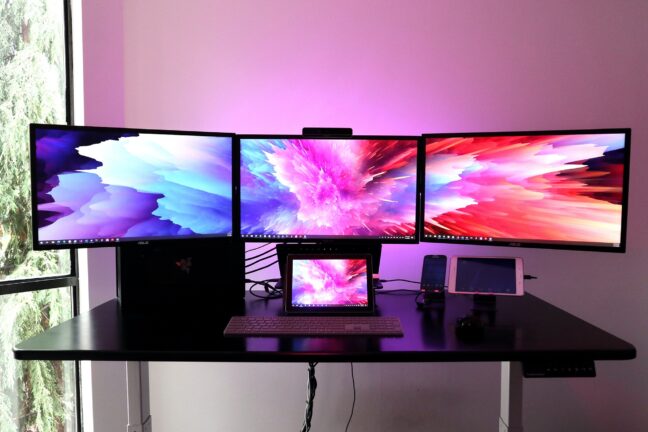 An office setup with three high-quality monitors, two tablets, a phone, and a keyboard in a dark room