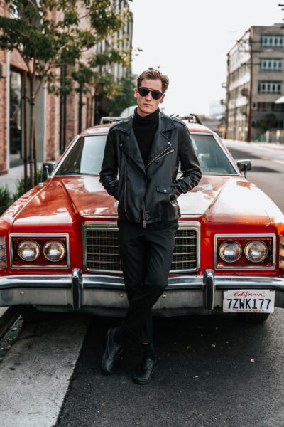 Man in black pants, a black leather jacket, and sunglasses leaning against a classic red car on a city street