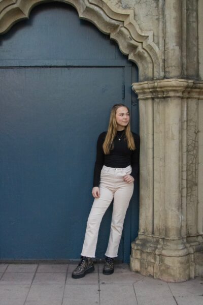 Woman in stylish boots, white pants, and a black top leaning against a pillar in front of a large blue door