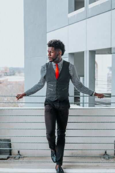 Full-body portrait of a man in a gray suit vest and nice pants leaning against a railing next to a building