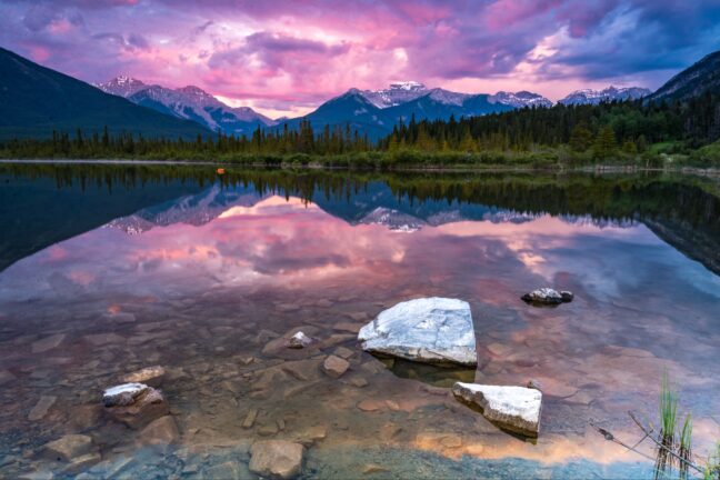 Lake with rocks in the foreground and mountains in the background under a purple sunset sky