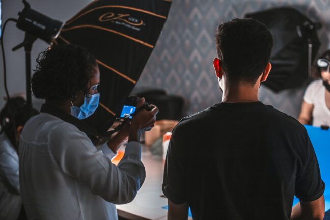 Photographer with blue mask holding up camera behind studio setup while people look on