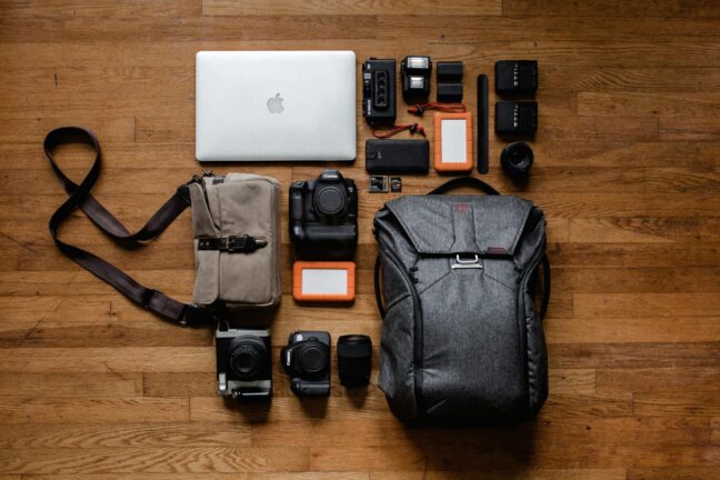 Camera backpack on a wooden floor surrounded by cameras, lenses, hard drives, and a laptop. 