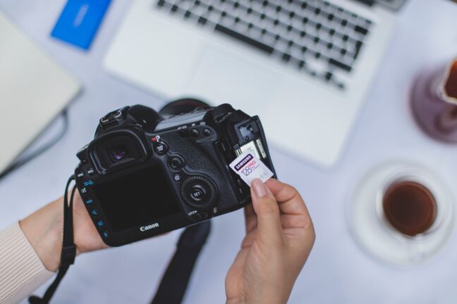 Hands taking a 128 GB memory card out of a black Canon camera with a laptop in the background.