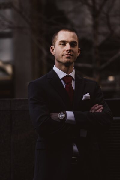 A man in a suit stands with crossed arms framed by a dark, moody background.