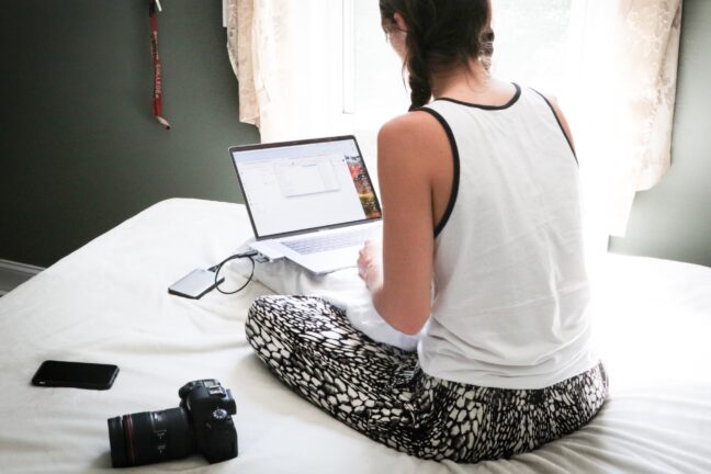 A woman sitting on a bed with a laptop and a hard drive plugged in with a camera at her side.