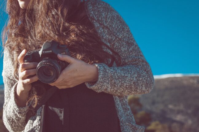 Woman holding a Canon EOS camera while wearing a gray sweater on a bright blue background.