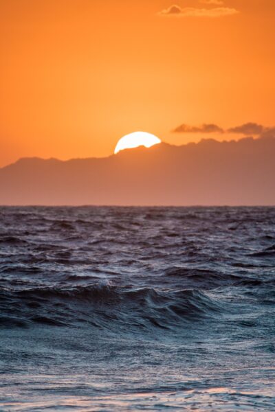 Orange sunset and disappearing sun with blue waves on a beach in the foreground.