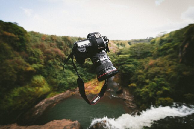 A black Canon DSLR tossed in the air with a waterfall, a pool, and trees in the background.