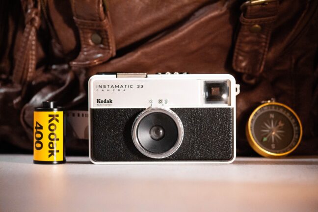 The white and black Kodak Instamatic 33 camera on a white surface next to a compass and a roll of film with a leather bag in the background.