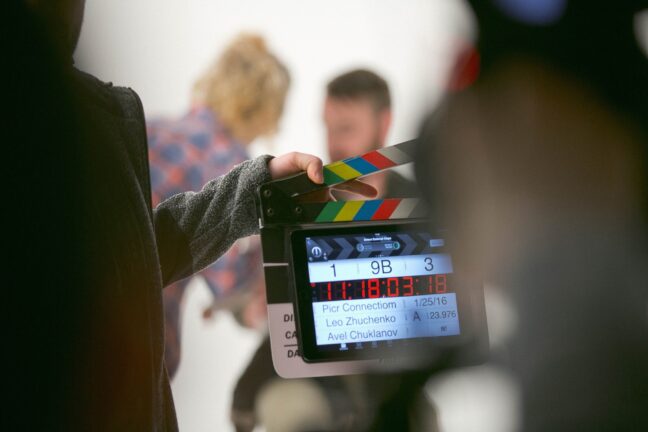 A hand holding a digital clapperboard in front of a blurry camera with people in the background.