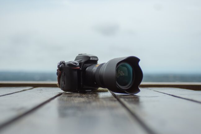 A black Nikon DSLR and a lens with a bulbous front element on a wooden dock with a blurry horizon in the background.