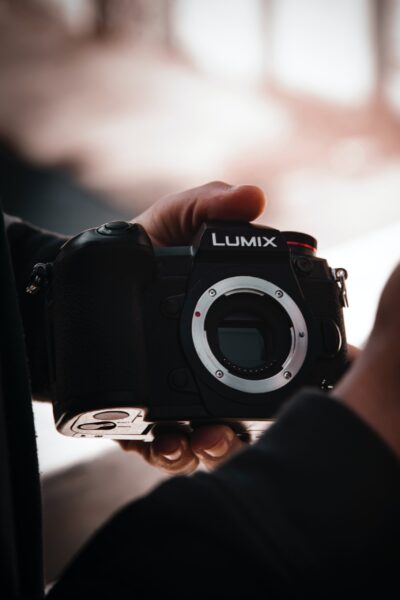 Hands holding a black Panasonic Lumix camera with the lens cap off and the sensor visible.