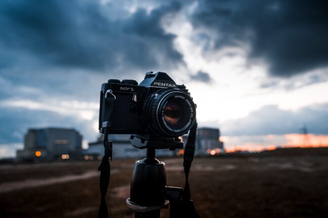 A black Pentax camera mounted on a tripod with dark sunset clouds and buildings in the blurry background.