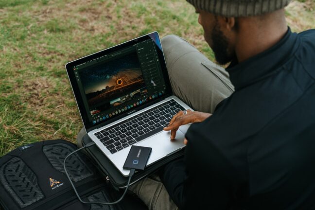 A man sitting on the grass while editing a fiery steel wool image on a laptop.