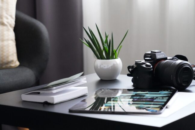 A black Sony mirrorless camera on a table next to a tablet, a journal, and a small potted plant.