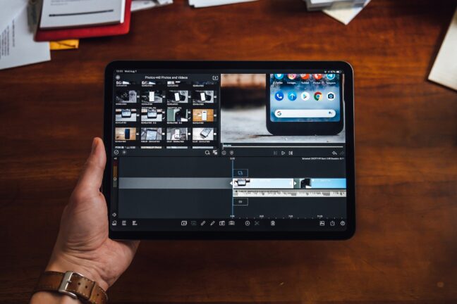 Hand holding a tablet above a desk with a video editing program showing thumbnails and recording files.