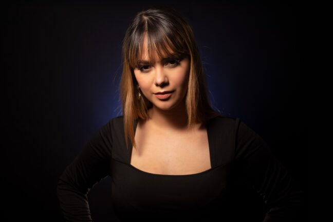 Woman with dark hair, red lipstick, and a black top looking at the camera with a faint blue light in the background.