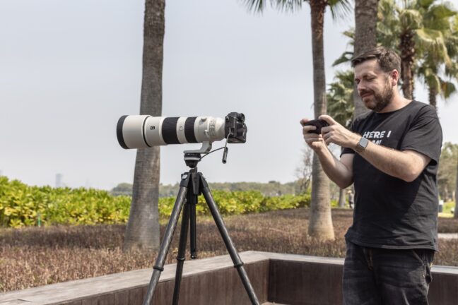 A man in the park using a smartphone to control the Benro Theta tripod with a camera and telephoto lens mounted.