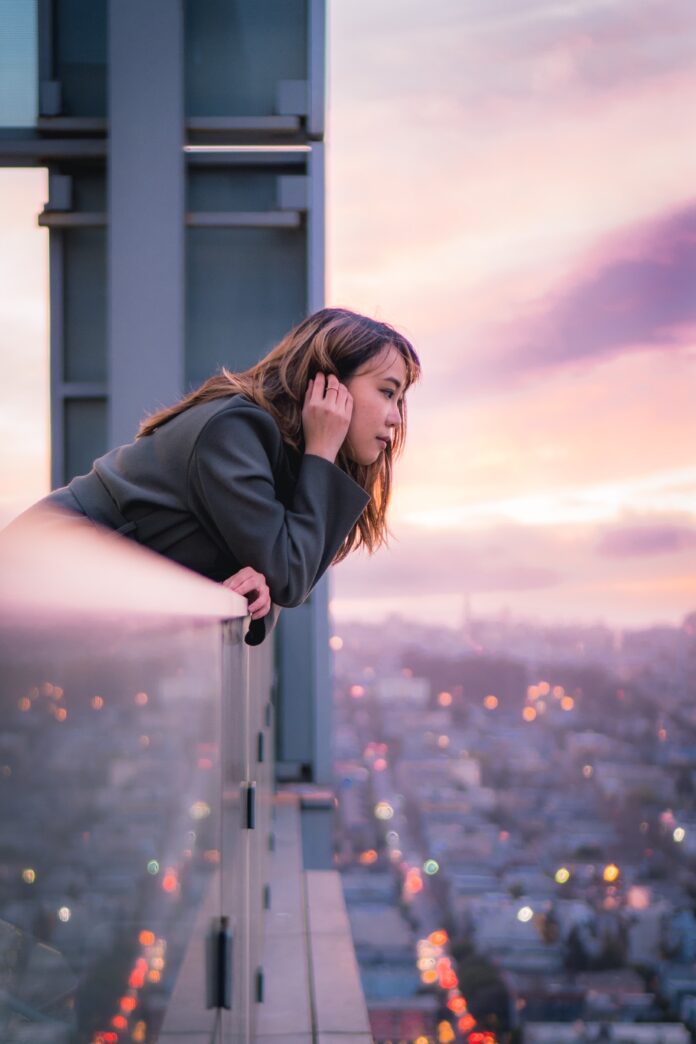 Woman leaning out over an apartment balcony many stories up above a city.