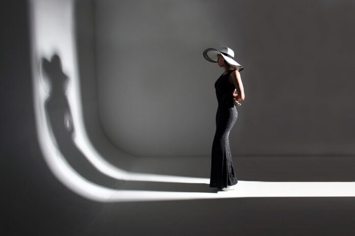 Fashion photoshoot with woman model wearing an elegant tight black dress and white hat facing her shadow on wall.