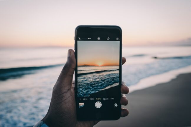 An iPhone held up to take a photo of the sunset over a beach.