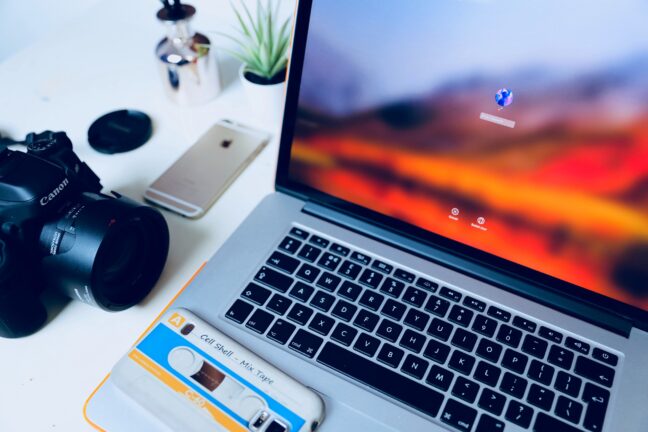 A Macbook Pro laptop displaying the login screen next to a black Canon camera and two smartphones.