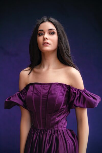 A dark-haired woman wearing a purple dress stands against a purple background.