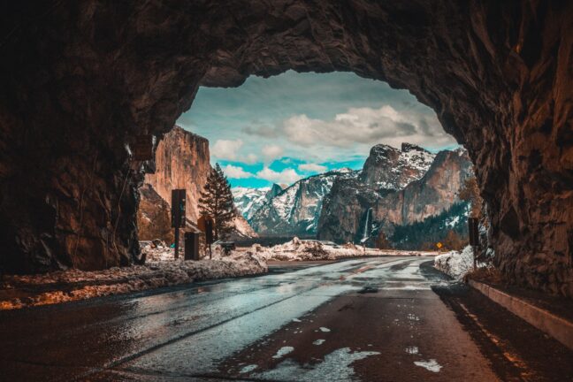 A rock tunnel with a road running through it frames mountains with a waterfall and a dramatic sky in the distance.