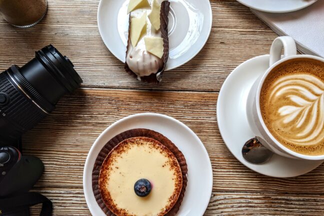 Coffee and desserts on white plates above a wooden table next to a camera.