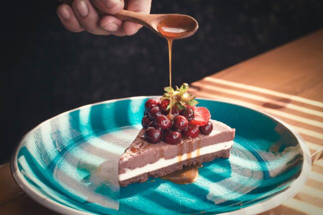 A hand holds a wooden spoon with caramel dripping from it onto a berry-covered cake.