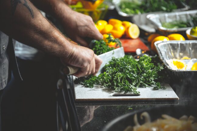 Hands using a large knife to cut parsley with lemons and vegetables in the background.