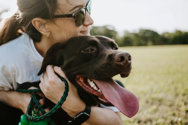 A young woman hugging a dark brown dog with its tongue out in a field.