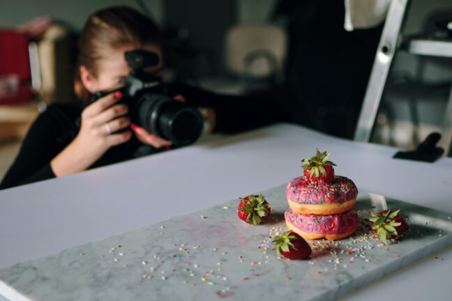 A photographer crouches next to a table while photographing donuts.