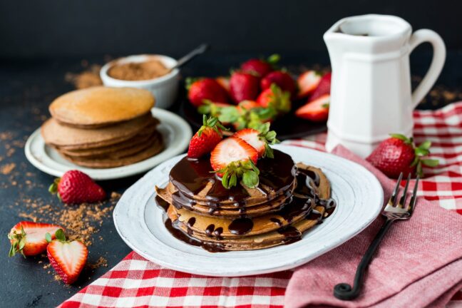 Pancakes with chocolate on a white plate with strawberries and cinnamon in the background.