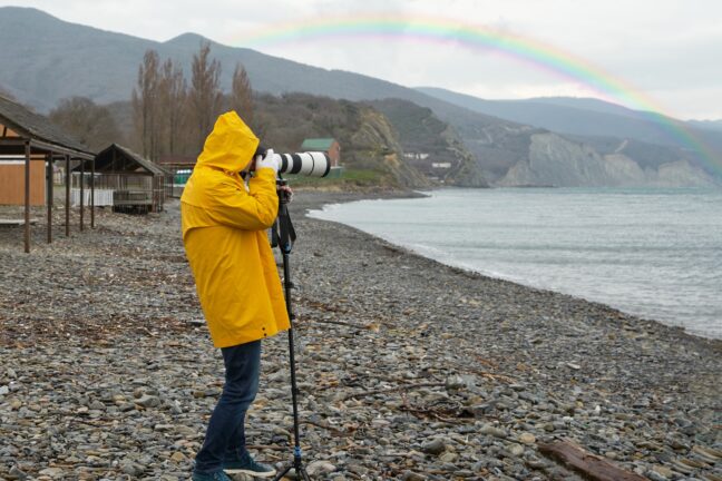 A man photographs a rainbow on the beach with a long-focus lens resting on a monopod.