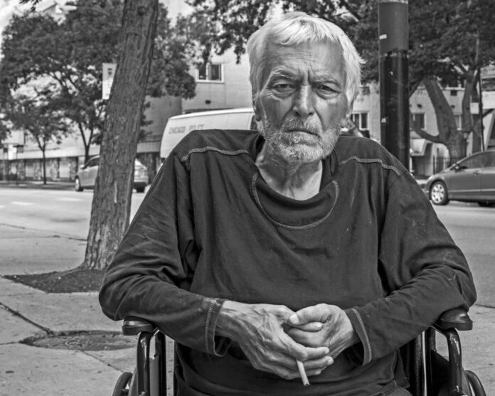 Man in a wheelchair holding a cigarette on a sidewalk next to road. 